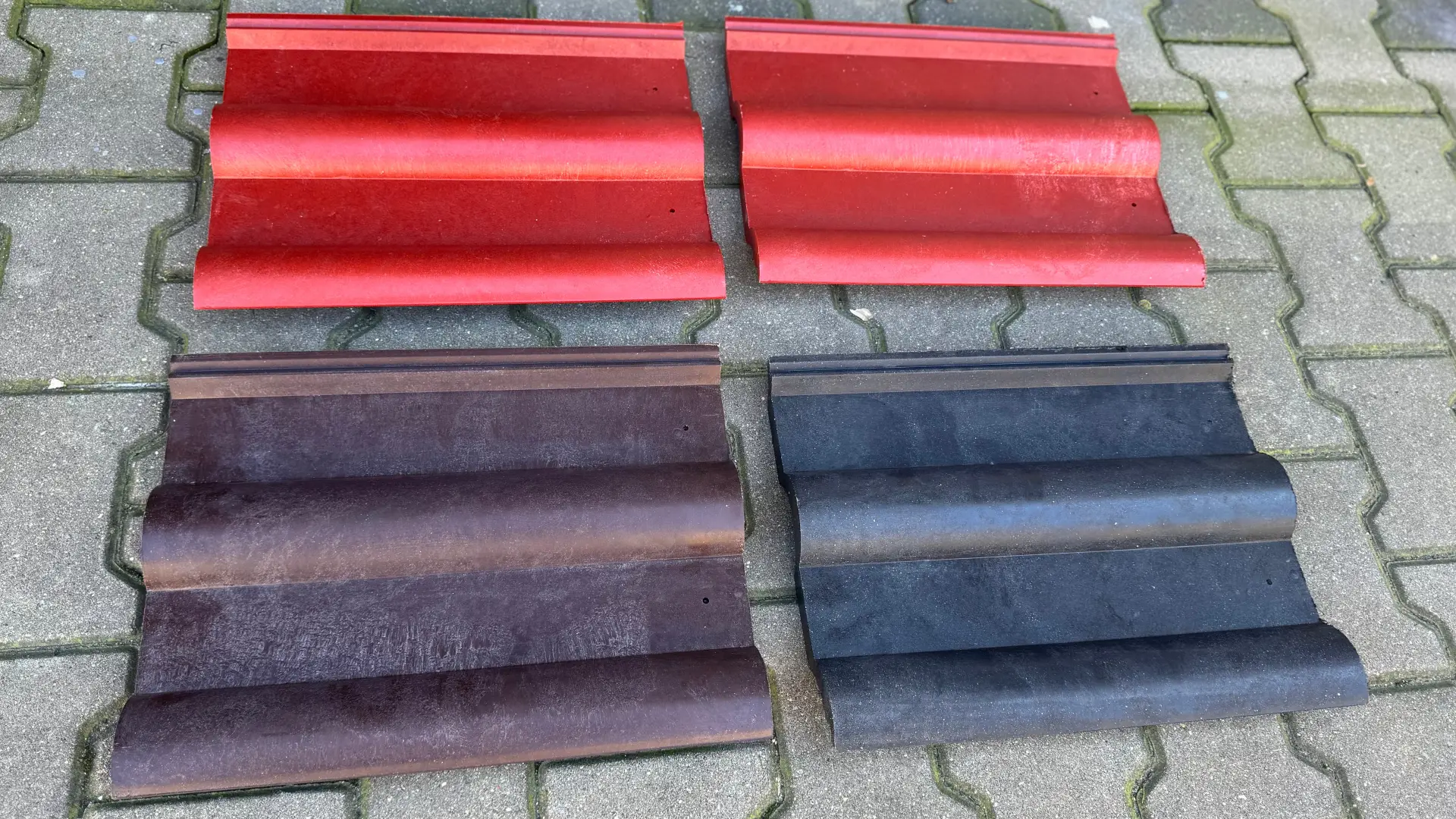 Four sets of colorful, composite roof tiles on a gray patterned pavement. The top two sets are red, while the lower sets are in shades of brown and black, organized neatly for display or selection.